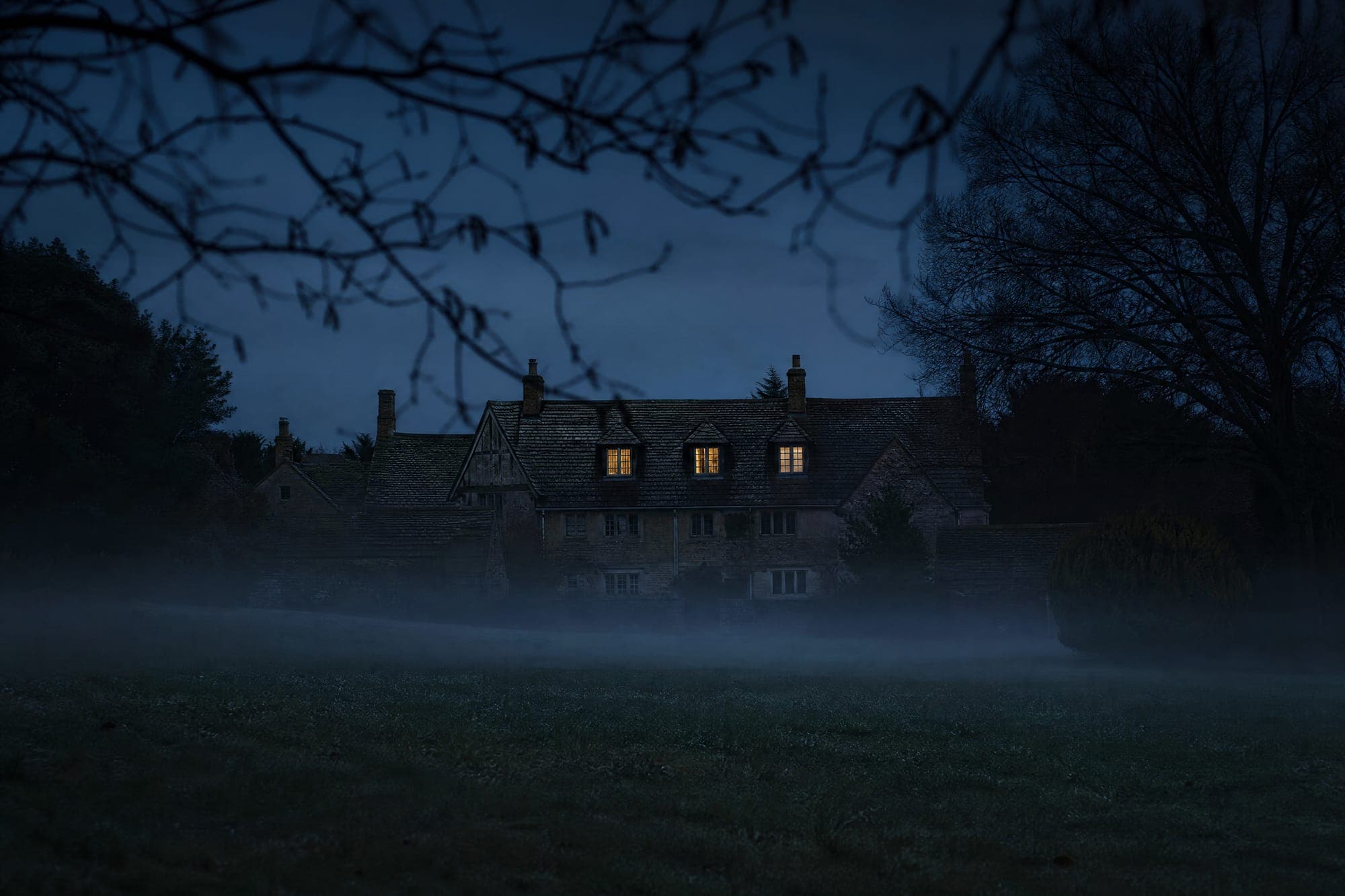 An old inn seen from across a dark lawn at dusk, three upstairs windows lit